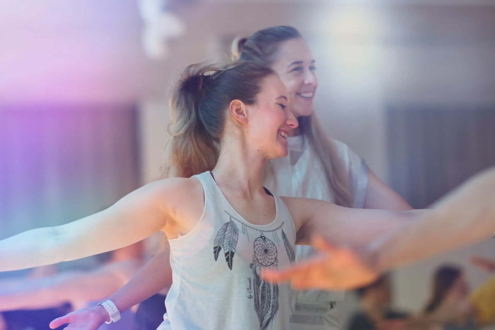 Zwei junge Frauen lachen mit ausgestreckten Armen im Studio, Dreamcatcher-Top, warmes Licht